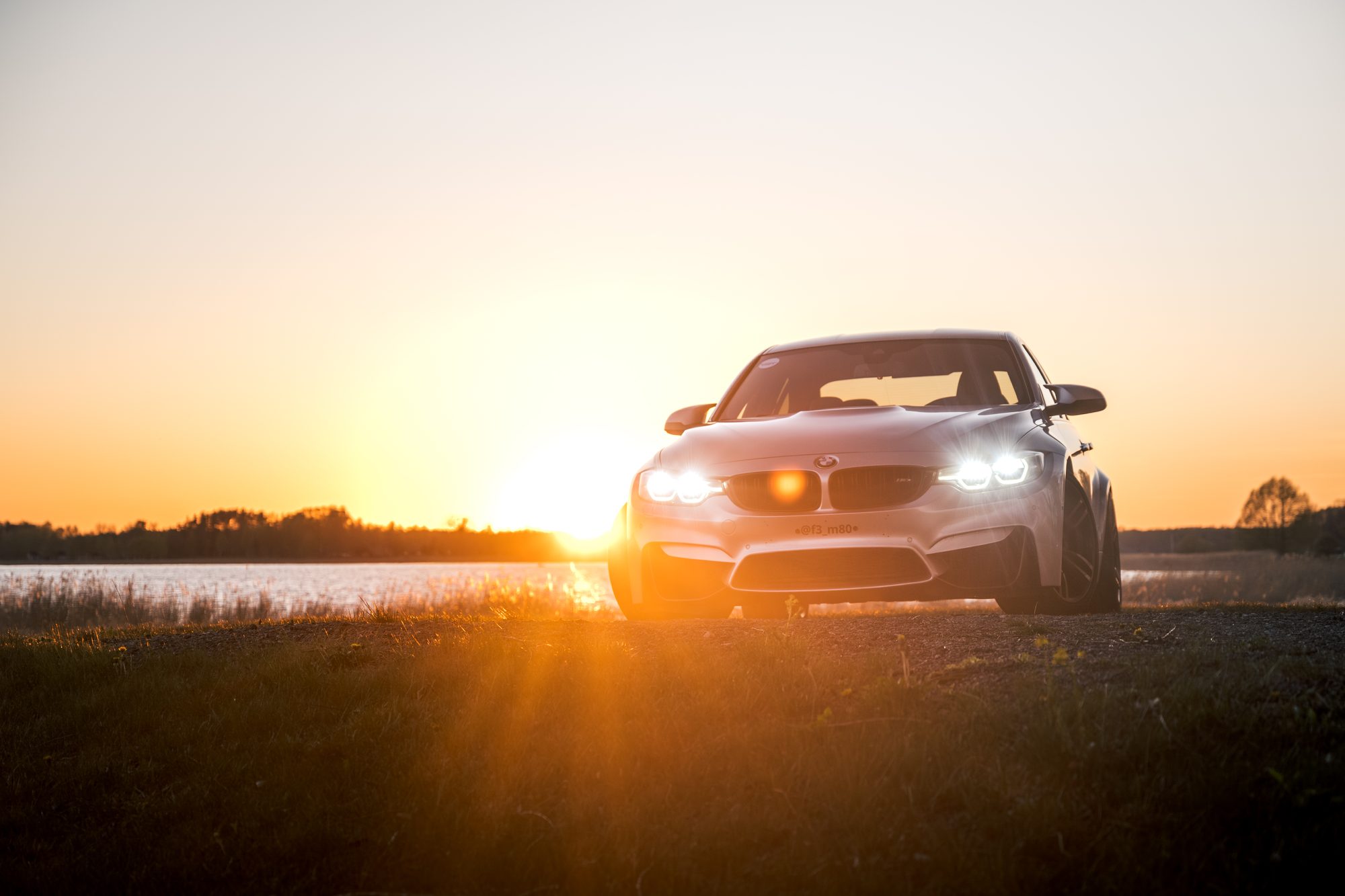 Silver BMW M3 F80 with its headlights on, the sun setting behind it over a lake